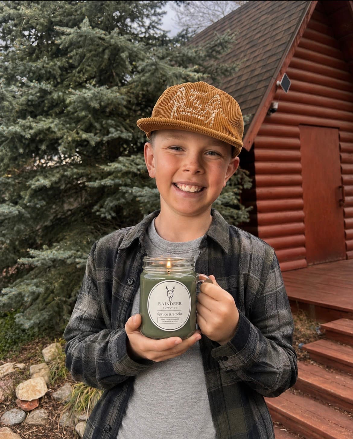 Rainier, one of the two young brothers behind Raindeer Supply Co, smiling and holding one of the family's hand-poured candles in front of a log cabin and pine tree.